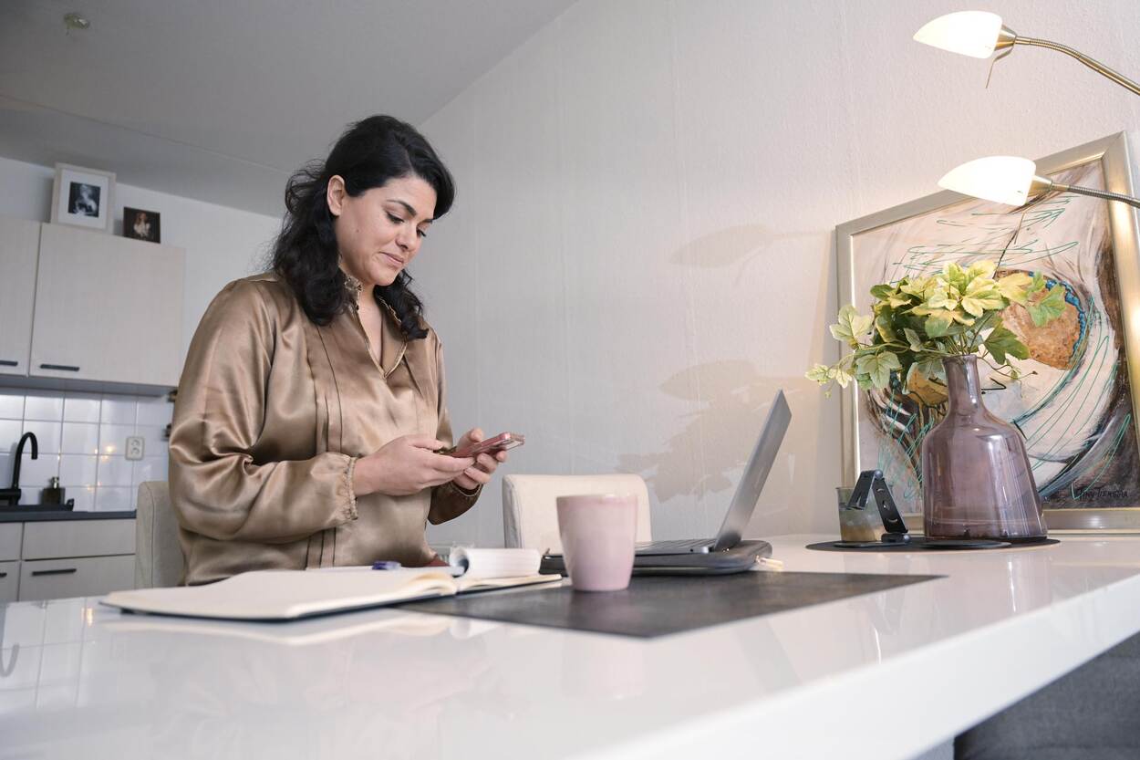 Een vrouw die met haar laptop en telefoon werk aan de keukentafel.