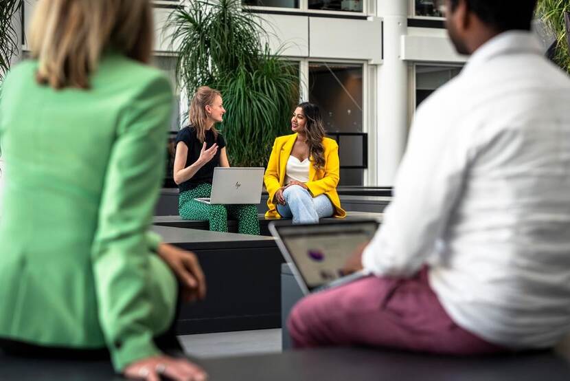 In het midden van de foto zitten twee vrouwen met elkaar te praten, waarvan er 1 een laptop op schoot heeft. Aan weerszijden van de foto zijn 2 mensen zichtbaar in een vergelijkbare situatie.