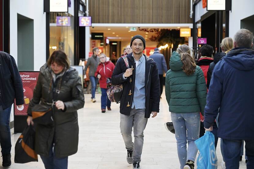 Een man met een glimlach op zijn gezicht en een tas op zijn rug loopt door een druk winkelcentrum.