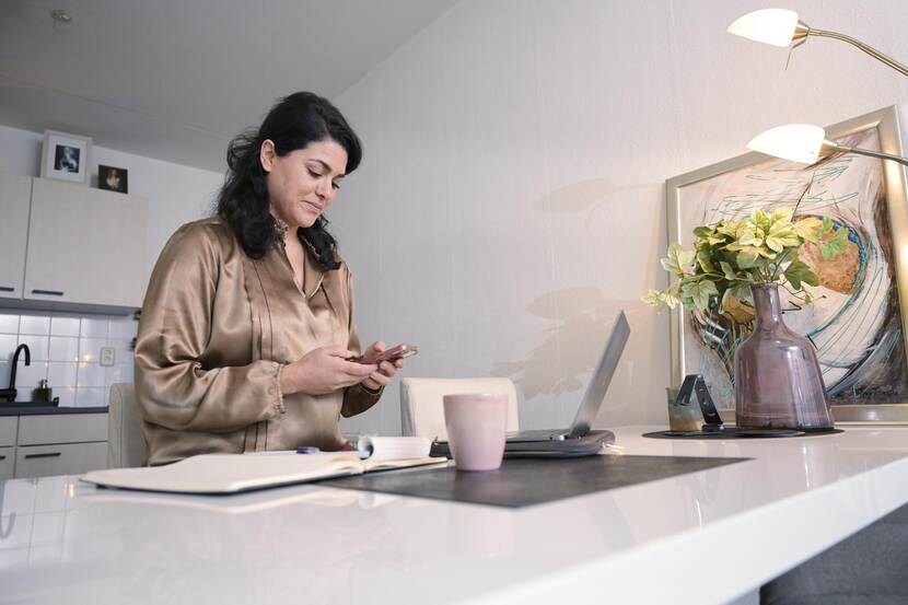 Een vrouw die met haar laptop en telefoon werk aan de keukentafel.